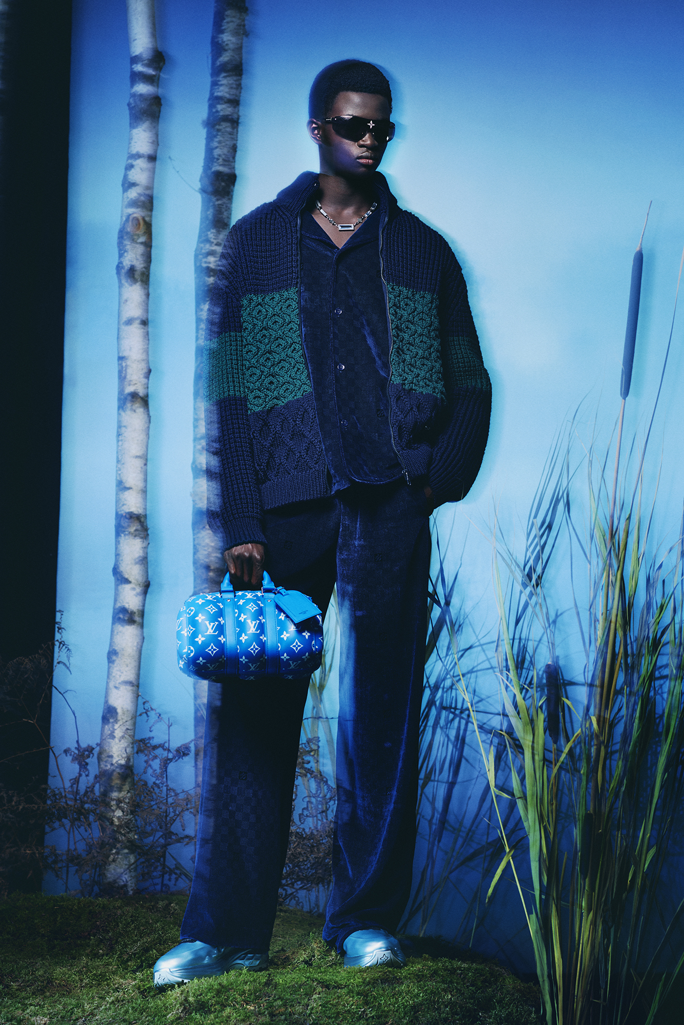 Men's Collection Pre Spring/Summer 2024 model with knit cardigan green and blue and teal-cyan handbag with dark pants by forest background