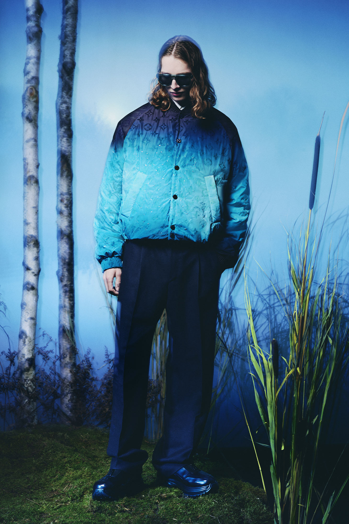Men's Collection Pre Spring/Summer 2024 model in cyan and blue varsity jacket and black pants and shoes in forest background
