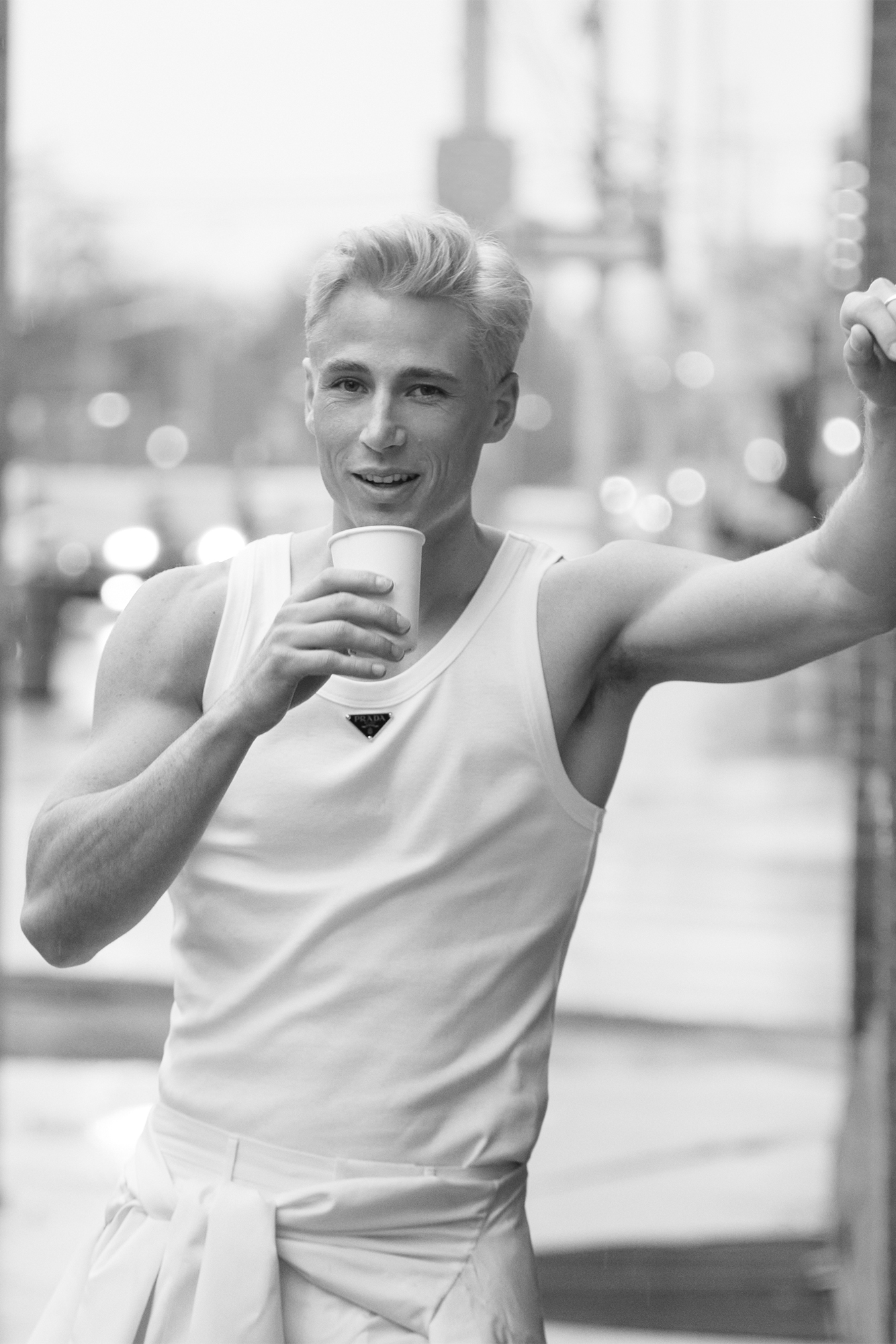 Male model in black and whiteholding drink and leaning against building in a white tank top
