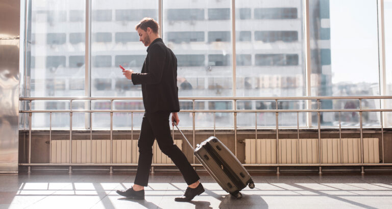 Carry On Luggage feature image. Man wheels suitcase in front of large airport windows on a clear day