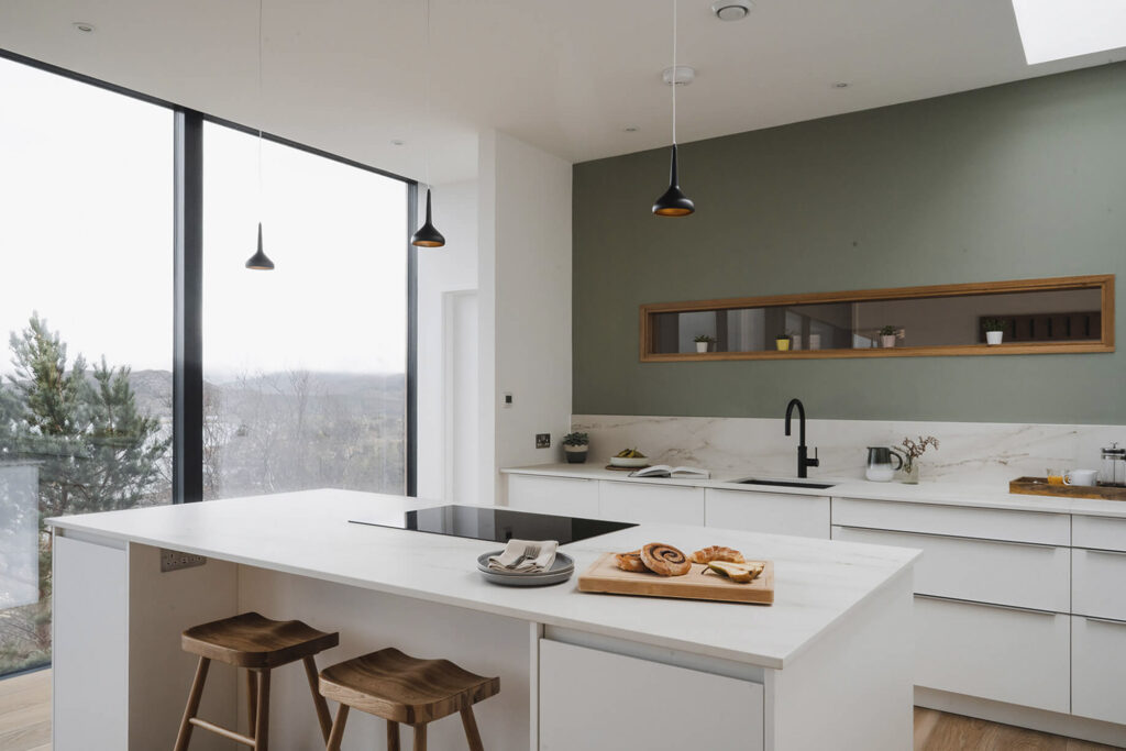 Kitchen with white countertops and green backsplash, big windows, and stools at kitchen bar