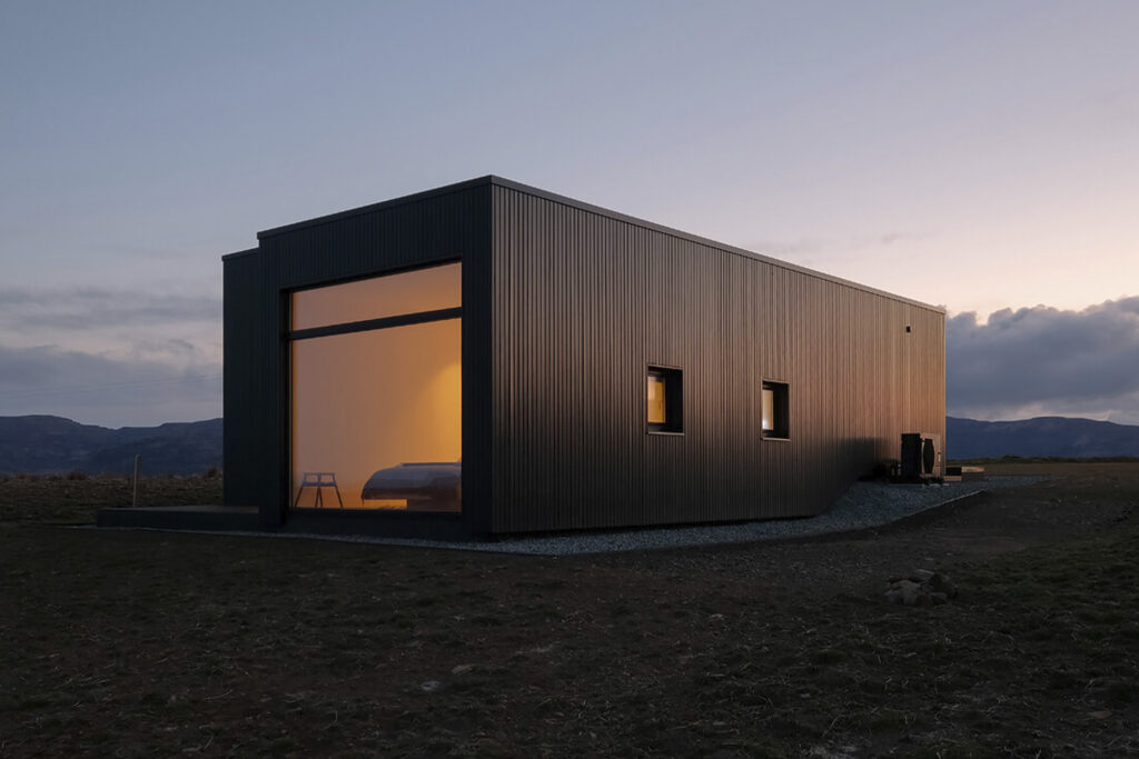 Harlosh Black H Home for rent in UK Scotland shot from outside at sunset. lit orange inside, black exterior