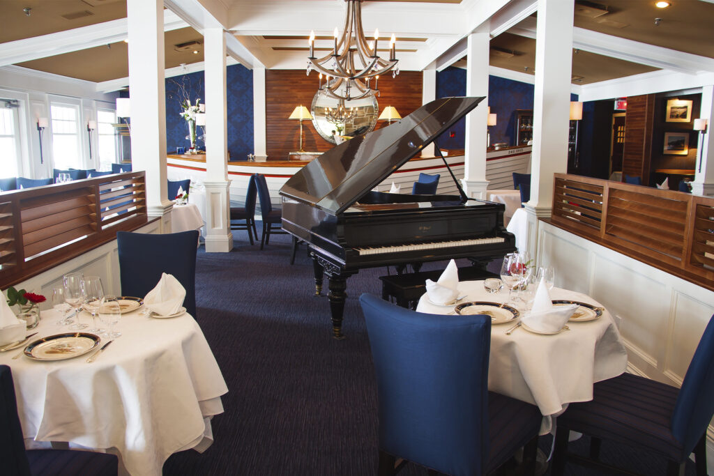 Le Riverain inside view grand piano with chandelier and white table clothes on tables with blue chairs