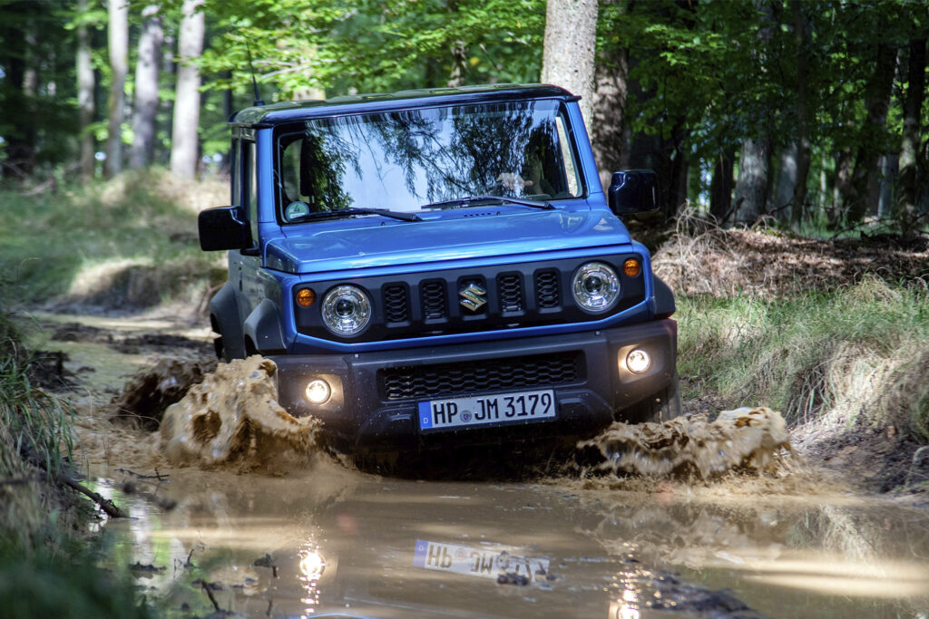 Blue Suzuki Jimny driving through muddy puddle in woods