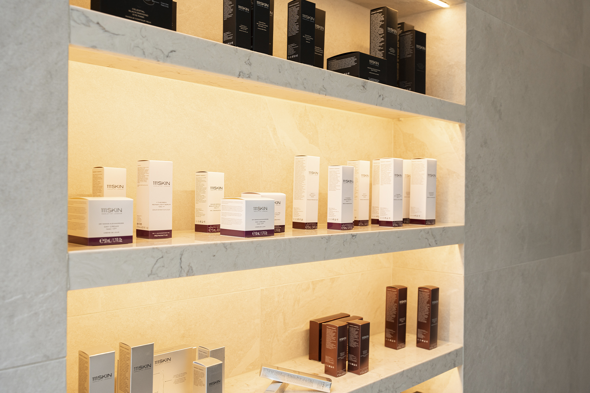 Shelf at Aman Spa Toronto showcases three shelves of skincare products: top shelf for has black containers, middle shelf has white containers, bottom shelf is white containers on left, brown containers on right.