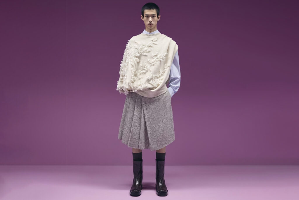 Model stands in front of purple background; he wears Dior Wellington Boots to show shows British Invasion in men's fashion, paired with a white dress top and shirt-kilt in grey