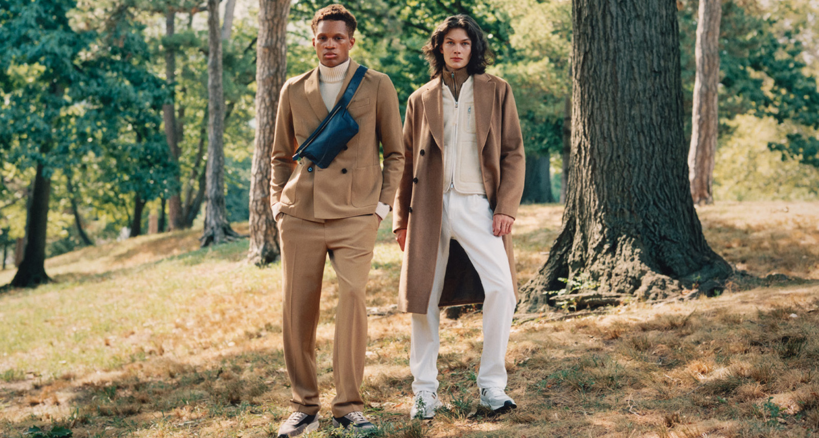 Two models in the woods; on left, the model is in a beige double-breasted suit, on right, the model is in a white shirt and pants with a beige open peacoat