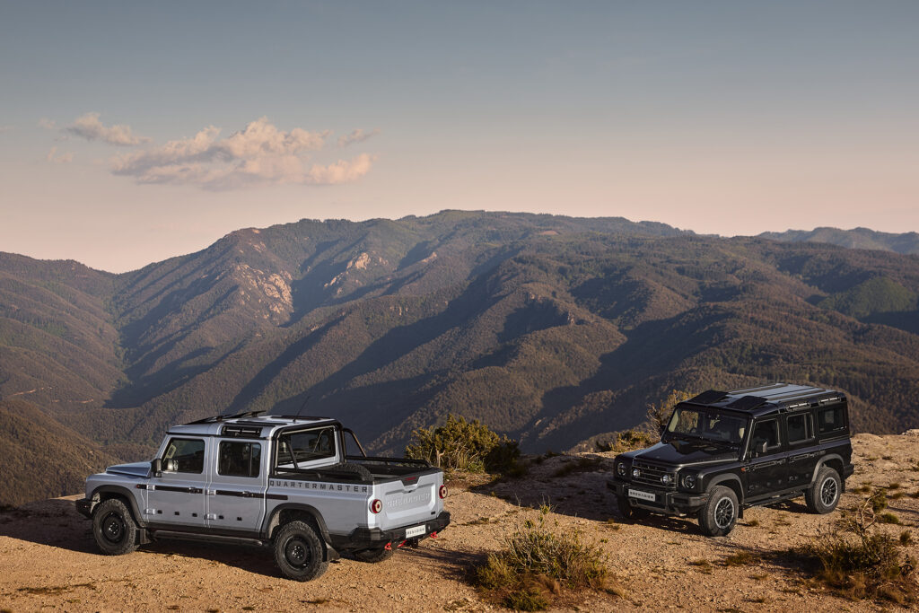 Two Ineos Grenadier Quartermaster, one silver and one black, parked on the top of a mountain showing a whole range in the background