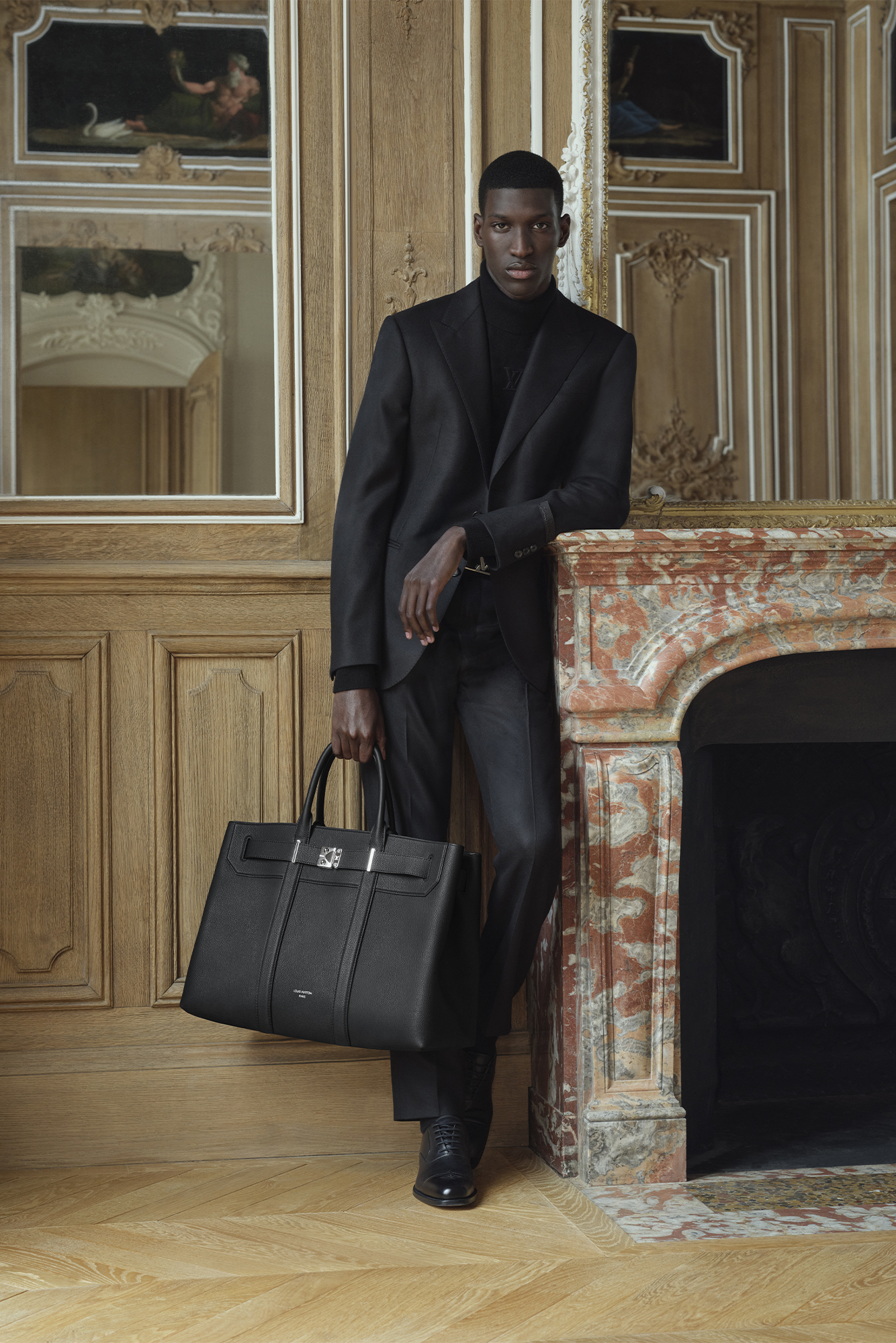 Model wearing a Louis Vuitton black outfit holds a bag and looks at camera while leaning by fireplace