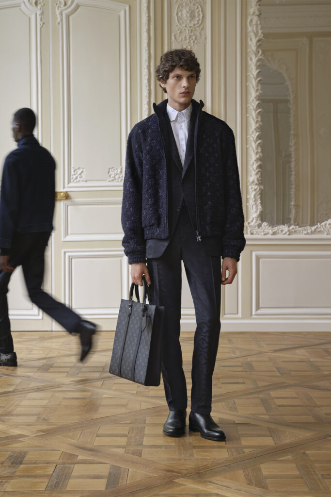 Model wearing a Louis Vuitton outfit and white shirt stands with bag