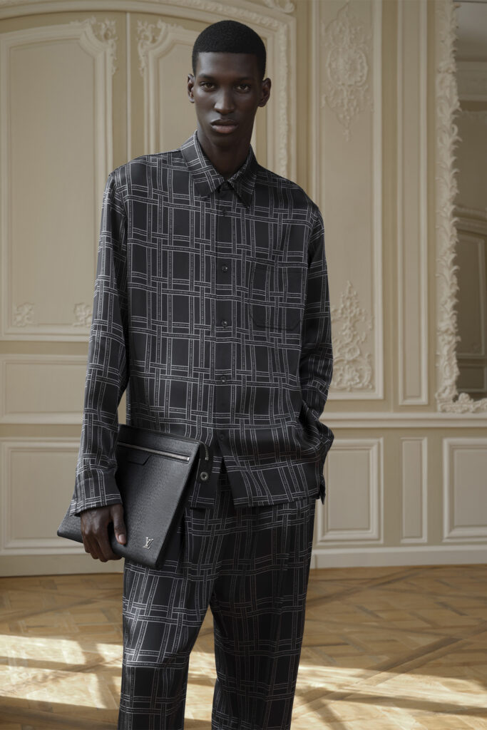 Model wearing a Louis Vuitton plaid patterned black outfit poses wile holding a dark case