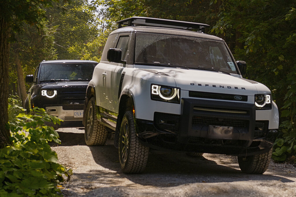 2 Land Rover Defenders drive off-road, in front is a white one, in back is a black one