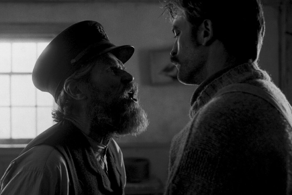 Willem Dafoe and Robert Pattinson look at each other in black and white photo with sun shining in through window