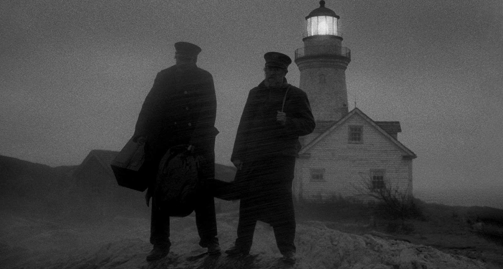 Two men on stand on rocks in front of a lighthouse on a grainy black and white photo
