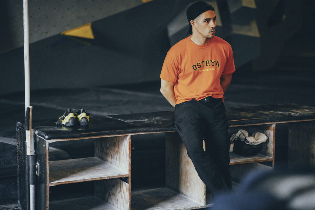 Ostrya founder Simon-David Fortin leaning up against shoe shelf in orange t-shirt, black pants and black hat