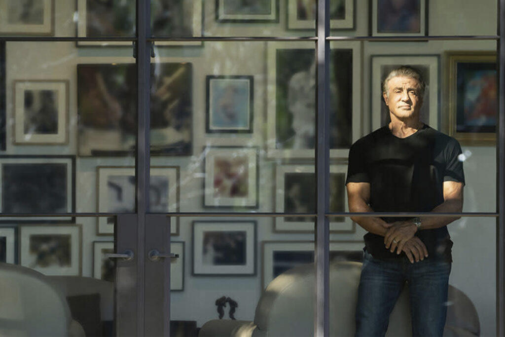 Sylvester Stallone stands in front of wall filled with frames for "sly"