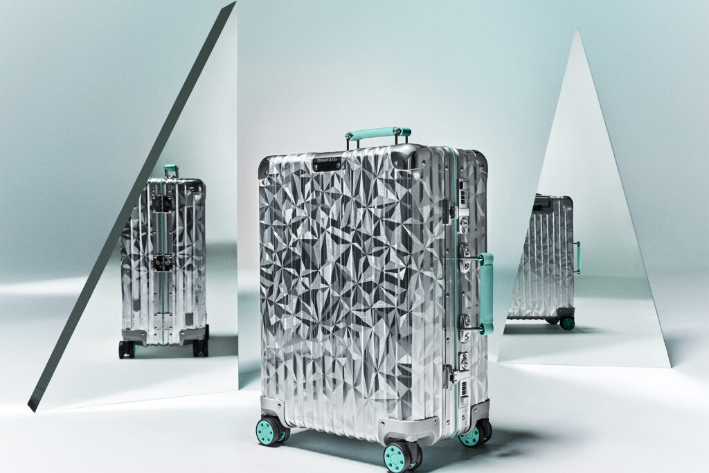 Shiny metal RIMOWA x Tiffany suitcase with signature blue-green wheels and handles and glass triangles in the background