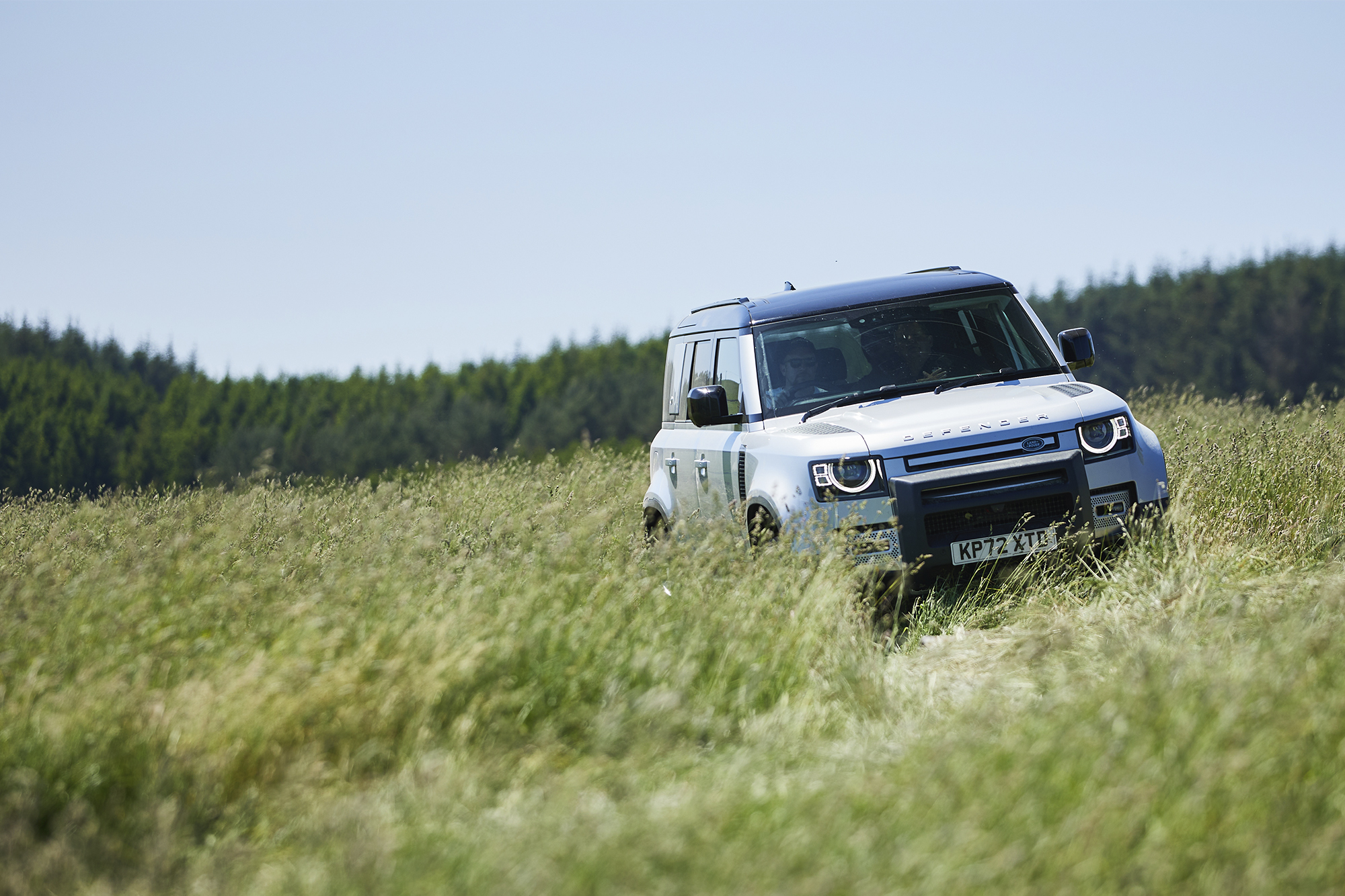 Land Rover Defender white, shot from the front, driving through a field on a sunny day