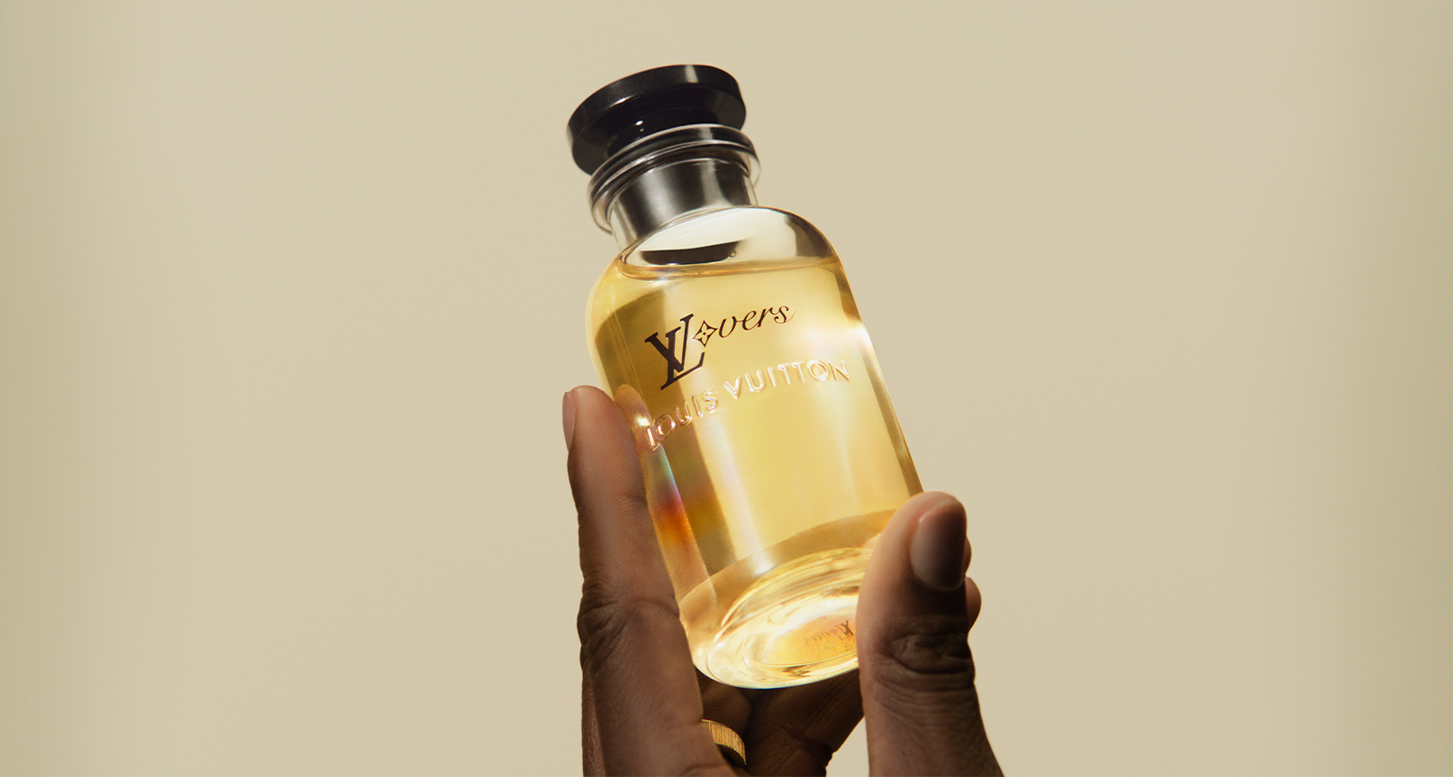 LVERS by Louis Vuitton Unisex Fragrance. campaign shot shows a hand holding a bottle from the bottom against a grey-yellow background. The bottle is filled with yellow fragrance