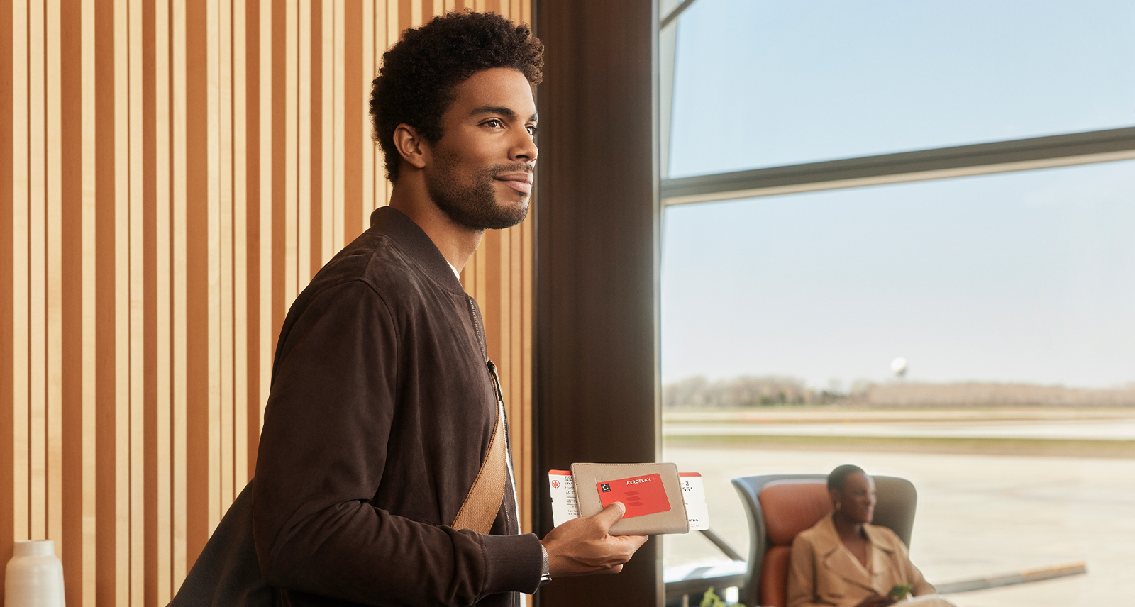 A man stands by an airport window. He's holding an Air Canada Aeroplan card.