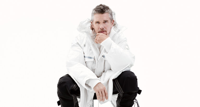 Ethan Hawke sits on a white background, wearing a white coat with black pants for Canada Goose