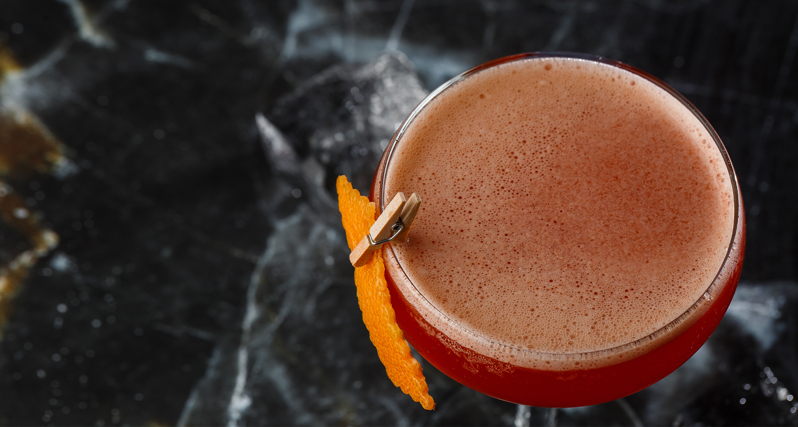 Photo shows a dark red nonalcoholic cocktail shot from above, with a citrus peel garnish pinned to the side.