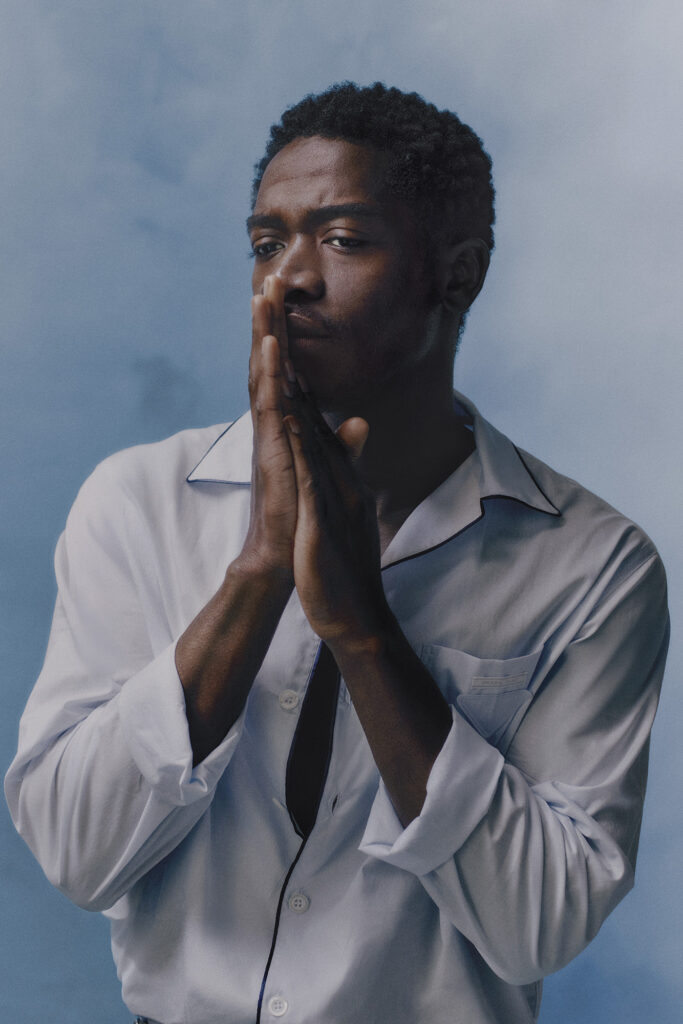 Damson idris shot from side with hands held up to his mouth. shirt by prada