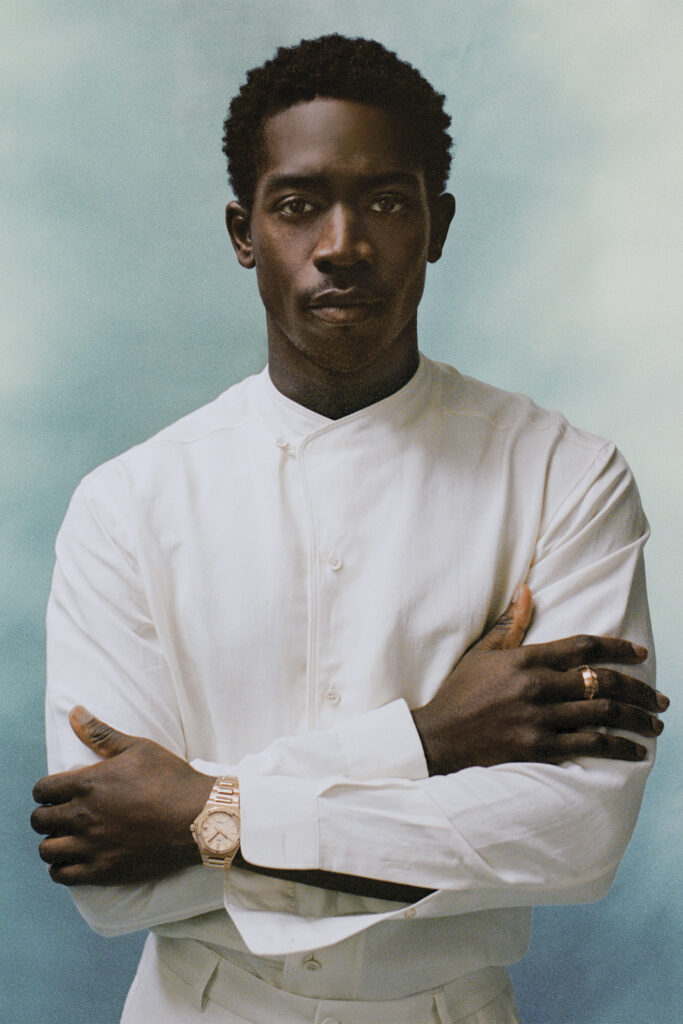 Damson Idris stand in from of blue background. Full look by loro piana, watch by iwc, ring by didris