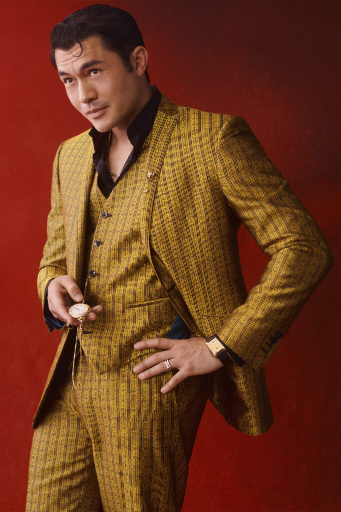 Henry Golding stands in front of a red background in a deep yellow suit.