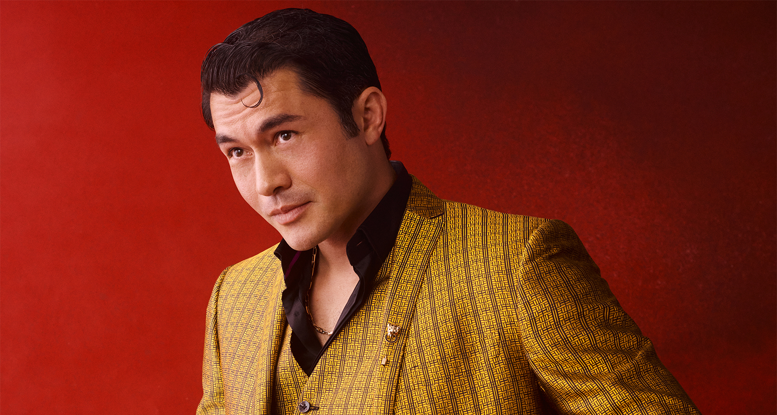 Henry Golding stands in front of a red background in a deep yellow suit.
