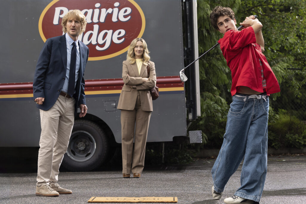 Peter Dager swings while owen wilson and a woman in a brown suit look on. Film still from "Stick."