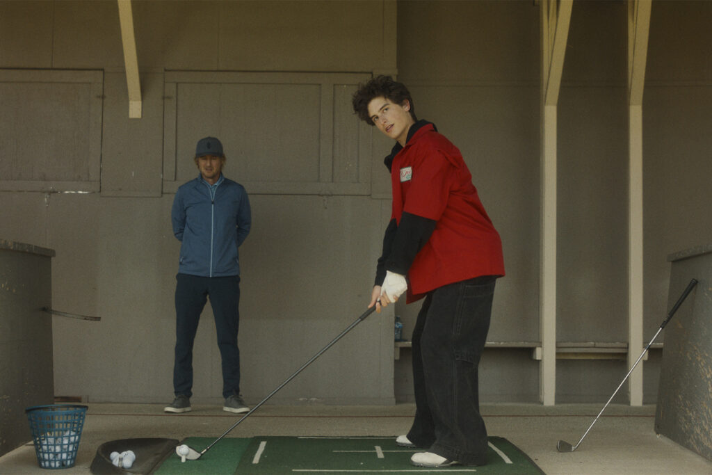 Photo still from "Stick" movie with Owen wilson standing in the bac while Peter Dager gets ready to hit a golf ball.