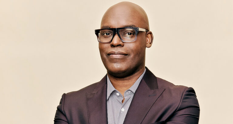 Cameron Bailey headshot on a yellow background. He's wearing black glasses and a brown blazer.