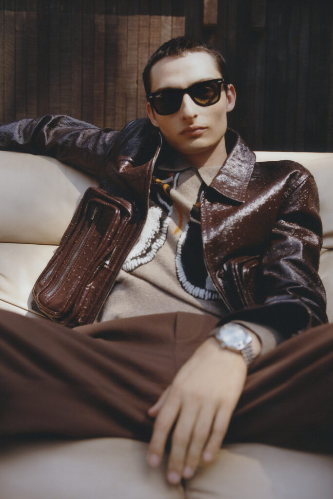 FINN WOLFHARD poses with sunglasses on, shot from below. ACKET, SWEATER, AND PANTS BY ACNE STUDIOS ; WATCH BY OMEGA ; SUNGLASSES BY RAY BAN ; COUCH COURTESY OF VISITOR GOODS