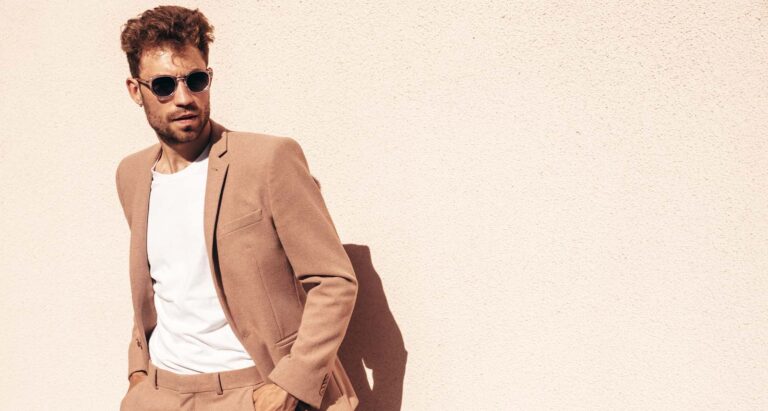 A man wears sunglasses, a white tee, and dust-orange blazer against a beige wall. Photo shows how men can dress during a heatwave.