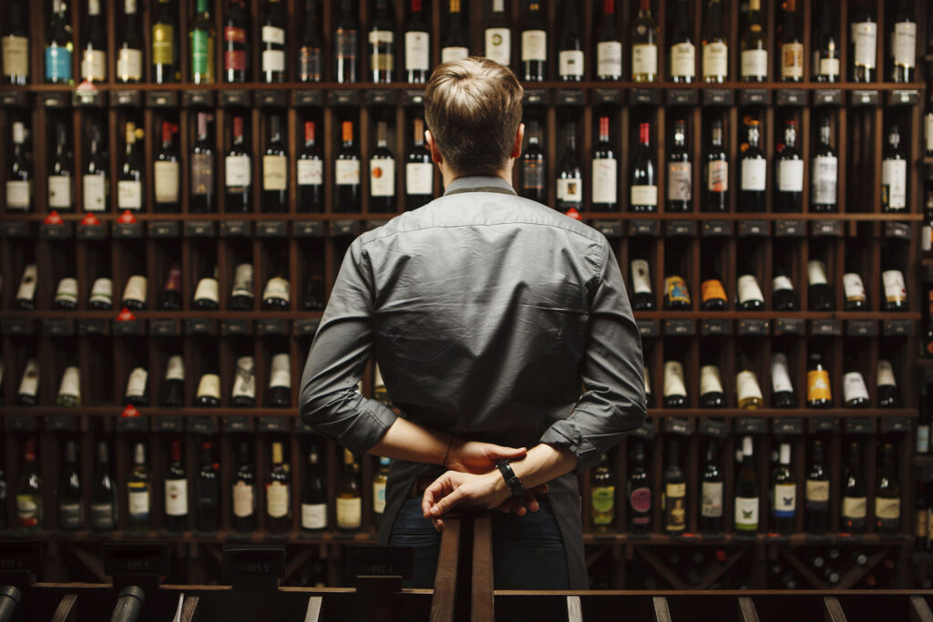 A man stands in front of a collection of vintage wines. Photo sourced from Adobe Stock.