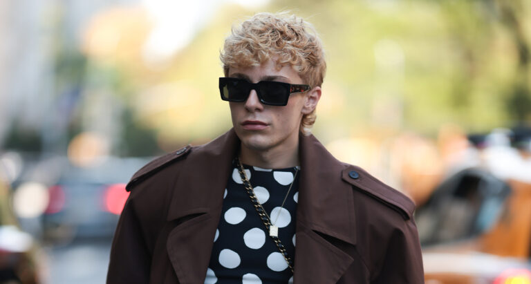 A man is seen with short, tousled blond hair and dark sunglasses. He wears a brown oversized trench-style jacket layered over a black shirt with bold white polka dots, paired with wide-leg black trousers. His look is completed with chunky black Rick Owens shoes and a statement chain necklace outside Vettese show during New York Fashionweek on September 14, 2025 in New York City. (Photo by Jeremy Moeller/Getty Images)