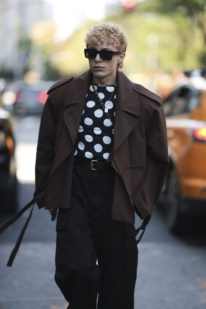 A man is seen with short, tousled blond hair and dark sunglasses. He wears a brown oversized trench-style jacket layered over a black shirt with bold white polka dots, paired with wide-leg black trousers. His look is completed with chunky black Rick Owens shoes and a statement chain necklace outside Vettese show during New York Fashionweek on September 14, 2025 in New York City. (Photo by Jeremy Moeller/Getty Images)