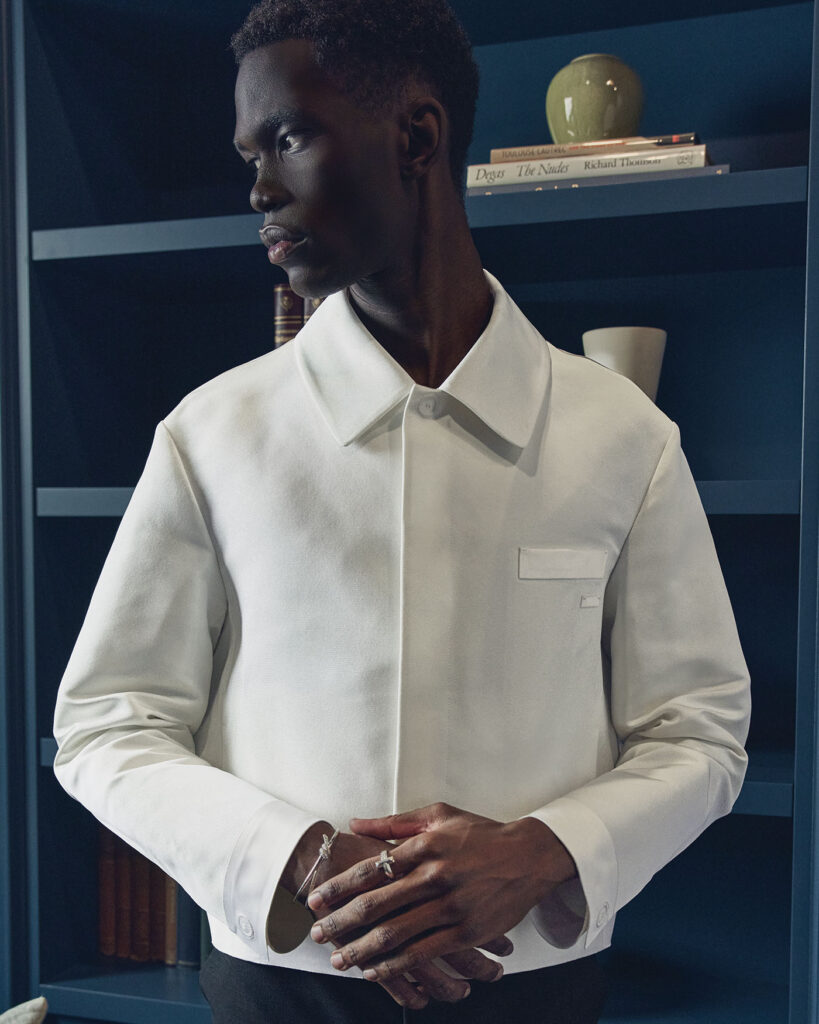 A male model wears a white jacket in front of a blue wall JACKET ($6,600) AND PANTS ($2,350) BY DIOR MEN; RING ($7,850) AND BRACELET ($41,200) BY TIFFANY & CO.