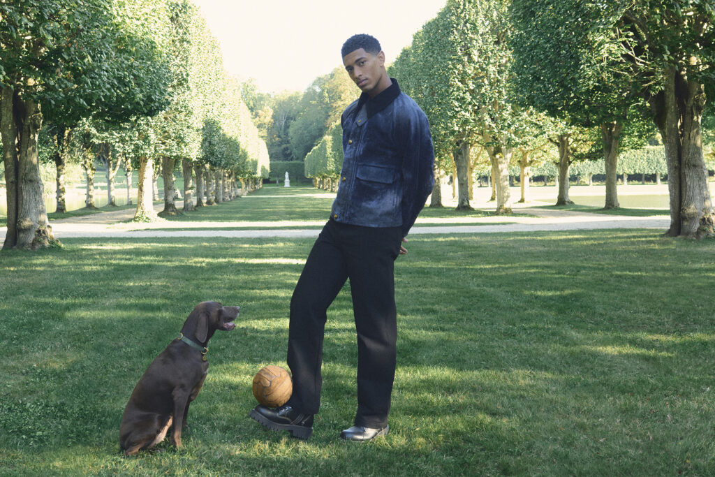 Model poses with chocolate lab in a field for LOUIS VUITTON MEN'S SS26 PRECOLLECTION DOG ACCESSORIES. PHOTO COURTESY OF LOUIS VUITTON.