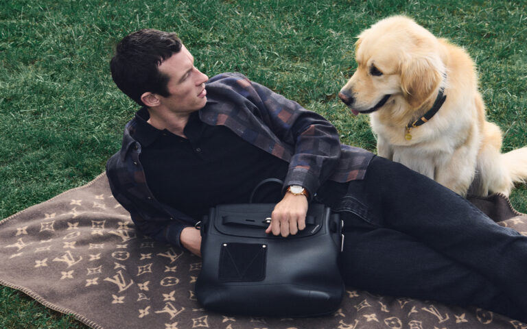 Callum Turner poses on a blanket in a field with a golden retriever for LOUIS VUITTON MEN'S SS26 PRECOLLECTION DOG ACCESSORIES. PHOTO COURTESY OF LOUIS VUITTON.