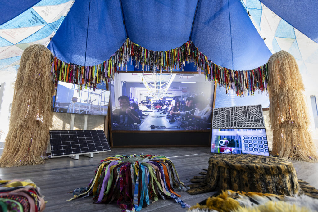 Exhibition view of “Dash” by Cao Fei Photo: Peng Jing Courtesy Fondazione Prada Cao Fei The Birth, 2026 Multimedia installation, variable dimensions A three-channel video loop, harvest props, an altar of worship, fertilizer bags, a banner, cushions, a XAG agricultural drone Structure of the installation developed by Small Production Banner and cushions designed by Xiang Gao Courtesy of the Artis, Vitamin Creative Space and Sprüth Magers Work produced by Fondazione Prada on the occasion of the exhibition “Dash”